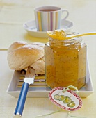 Gooseberry and juniper jam in open jar with bread roll and coffee on plate