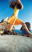 Frau im gelber Sommerkleidung am Meer, springt in die Luft, hält Ball