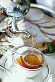 Aus einer Silberkanne wird Tee durch ein Sieb in eine Tasse gegossen