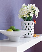 weiße Blumenvase mit Narzissen, Obstschale auf weißem Bord