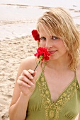 Portrait of beautiful woman wearing green top standing on beach and smelling red carnation