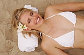 Pretty woman in white bikini with flower in hair lying on sand, smiling with eyes closed