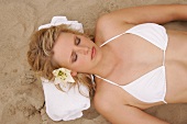 Pretty woman wearing white bikini lying on sand with towel under her head, eyes closed