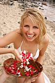 Pretty blonde woman sitting and holding bowl of white and red petals on beach, smiling