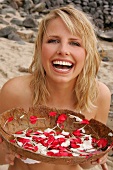 Pretty blonde woman sitting and holding bowl of white and red petals on beach, smiling