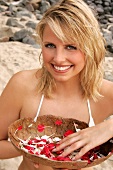 Portrait of beautiful blonde woman holding bowl of colourful petals on the beach, smiling