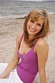 Portrait of beautiful blonde woman wearing purple top sitting on beach, smiling