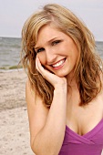 Portrait of pretty blonde woman in pink top with hand on face standing on beach, smiling