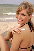 Close-up of attractive blonde woman sitting on beach and applying cream on back