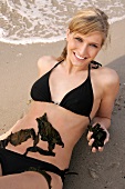 Portrait of pretty woman in black bikini lying on beach with seaweed on her belly, smiling