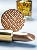 Close-up of gold cosmetic products, lipstick, eye shadow with nail polish in background
