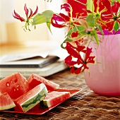 Melonenstücke auf einem Teller neben einer Blume auf einem Rattantisch.