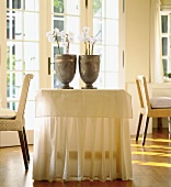 Tisch mit langer Tischdecke, darauf 2 Krüge mit Pflanzen, heller Raum