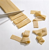 Pasta dough being sliced in wide strips with knife on white background