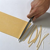 Hand cutting pasta dough into strips with knife on white background