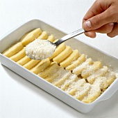 Parmesan being sprinkled on gnocchi discs in baking dish