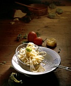 Pasta in strainer on plate besides tomatoes, parmesan, basil and herbs on wood