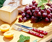 Bread with cheese, grapes, halved peach and cutlery on wooden chopping board