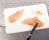 Removing tendons and fat from chicken breast on chopping board