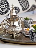 Close-up of silver tray with silver teapot, black-white glass and muffins
