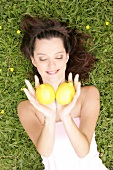 Pretty woman lying on meadow with eyes closed holding two lemons in cupped hands, smiling
