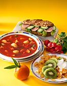 Close-up of vegetable soup, cornflakes with kiwi, sandwich, orange and radishes