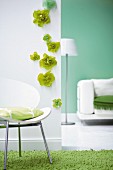 Weisser Stuhl mit Kissen vor weisser Wand mit grünen Papierblüten, daneben Spiegel