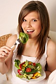 Portrait of woman with long hair holding bowl of salad and eating cucumber, smiling