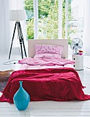 Bett mit Bettwäsche in Rottönen und japanischen Motiven, zwischen Stehlampe mit weißem Schirm und zylindrischem Nachttisch, vor Terrassenfenster mit Gartenblick