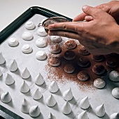 Close-up of hand sprinkling cocoa on decorative meringue on baking tray, step 3