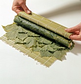 Close-up of hand rolling bamboo mat stuffed with rice on grape leaves, step 2