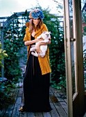 Frau mit auffälligem Hut und langem Kleid steht draußen, Katze auf Arm