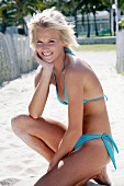 Beautiful blonde woman in light blue bikini sitting on sand with head on hand, smiling