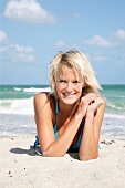 Portrait of beautiful blonde woman wearing blue bikini lying on beach, smiling