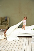 Cute child standing on bed and pulling white bedsheet