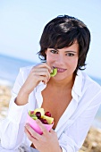 Brunette woman wearing white shirt sitting on beach while having fruit from bowl