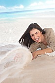 Ecstatic woman wearing beige sweater lying and in the sand, laughing