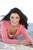 Portrait of pretty woman with windswept hair wearing pink sweater lying on sand, smiling