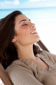 Woman wearing beige top relaxing on deckchair at beach, eyes closed