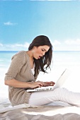 Side view of pretty woman wearing brown cardigan working on laptop while sitting on beach