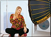 A blond woman in a flowery blouse sits on a table and points at the camera