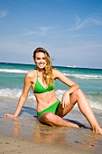 Portrait of happy blonde woman in green bikini sitting on beach, smiling