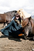 Beautiful brown haired woman wearing poncho and scarf crouching next to iceland horses