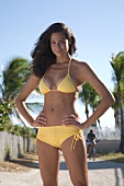 Woman with dark hair wearing yellow bikini standing on beach with hands on hips