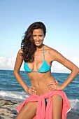 Woman with long hair in blue bikini, standing with hands on hip, smiling on beach