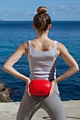 Rear view of woman holding ball over head while exercising for shoulder and chest near sea