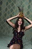 Portrait of happy woman wearing polka dotted dress holding pineapple on her head, laughing
