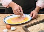 Close-up of hands dipping meat in egg yolk for preparation of schnitzel, step 2