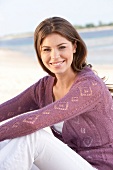 Portrait of beautiful brunette woman wearing purple sweater sitting on beach, smiling