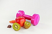 Halved kiwi, piece of chocolate, apple, carrot and pink dumbbell on white background